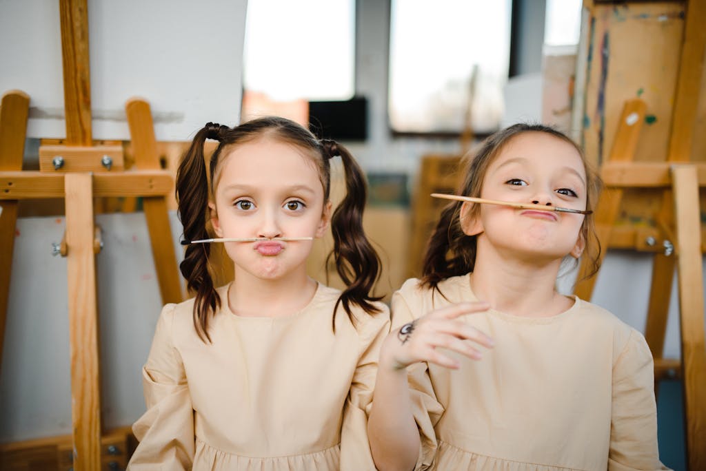 Two young girls making funny faces with brushes in an art class setting.